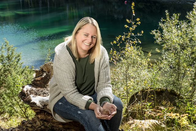 Nicole Gansner sitzt lachend am See und hält ein Schneckenhaus